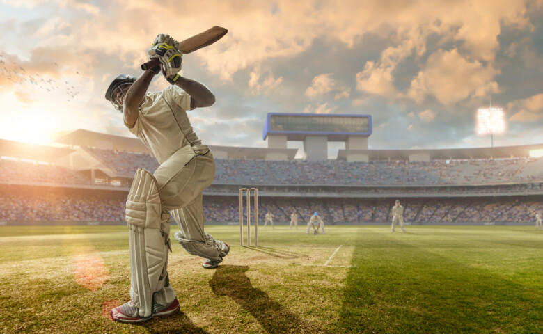 A close up image of a professional cricketer playing in batsman position wearing cricket whites and safety helmet, having just hit a ball during a cricket match in an outdoor stadium full of spectators. The action occurs under an evening sky at sunset. Stadium is fake, created from photographic and CG elements.